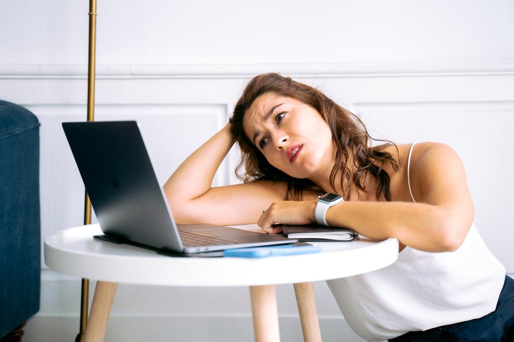 A frustrated woman sits at a desk with her hand on her head, staring at an open laptop.